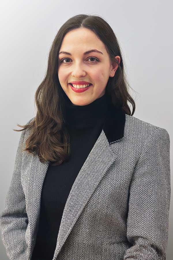 Employee photo by Nicole Lemke. A woman in a black sweater and a grey blazer stands in front of a white wall.