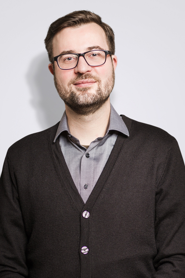 Staff photo of Sven Kurzke. He wears black glasses, a gray cardigan and stands in front of a white wall.