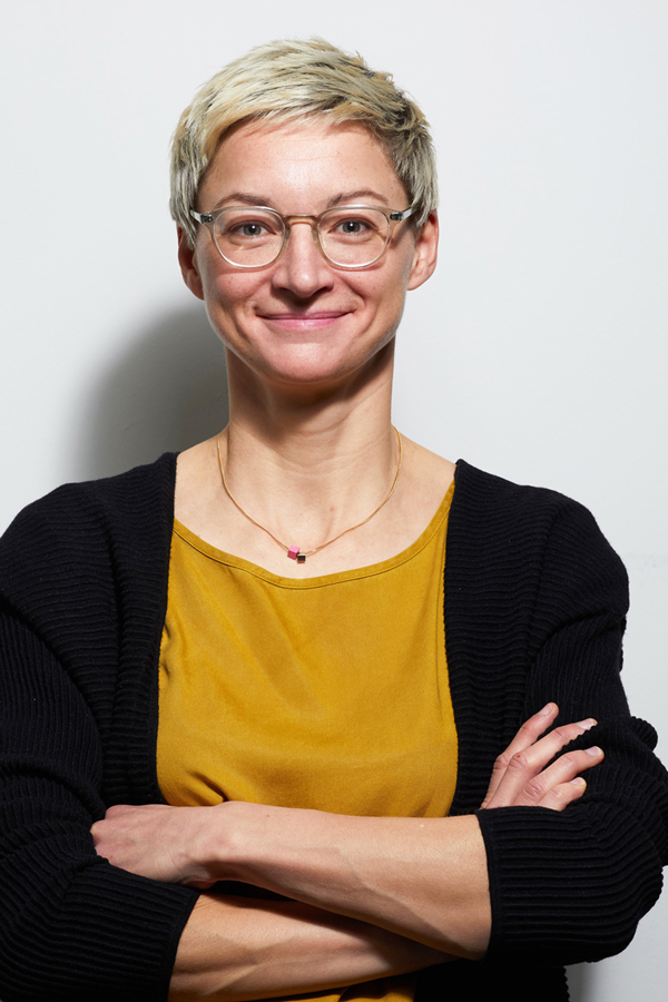 Employee photo of Wera Mohns Patten. She is wearing a yellow T-shirt and black cardigan and is standing in front of a white wall.
